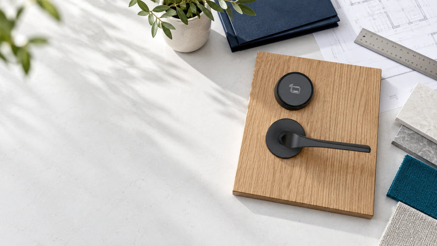 Matte black lever and circular reader on an oak sample panel beside architectural paperwork and material swatches.